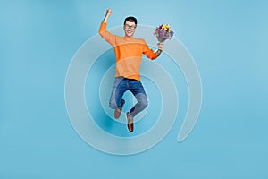 Full size photo of astonished handsome guy jumping fist up hold flowers isolated on blue color background