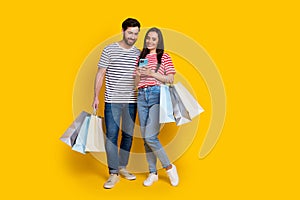 Full size photo of cheerful funny guy lovely nice girl look at smartphone holding shopping bags isolated on yellow color