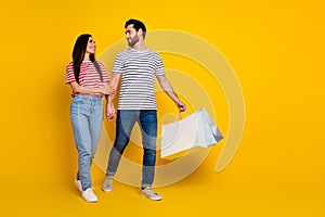 Full size photo of cheerful man look at girlfriend hold shoppings bags go to empty space isolated on vibrant yellow