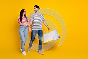 Full size photo of cheerful man look at girlfriend hold shoppings bags go to empty space isolated on vibrant yellow photo
