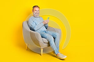 Full size photo of cheerful man sit chair hold use telephone chatting isolated on yellow color background