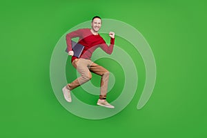 Full size photo of cheerful pleasant guy wear red shirt brown trousers running holding laptop in hands  on green