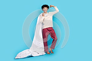 Full size photo of cute young guy hold blanket raise fist flexing power wear trendy white pajama outfit isolated on blue