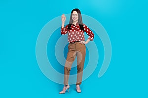Full size photo of friendly person wear red blouse brown trousers waving palm say hi arm on waist  on blue color