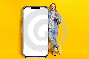 Full size photo of handsome cheerful man wear jacket jeans trousers lean on touchscreen smartphone  on yellow