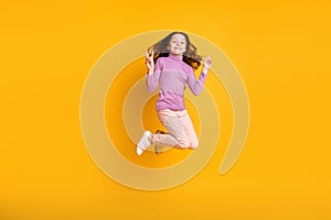 Full size photo of happy excited smiling cheerful little girl jumping showing v-sign  on yellow color background