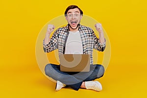 Full size photo of a happy young man holding laptop computer sit on a floor rejoice victory isolated over yellow color