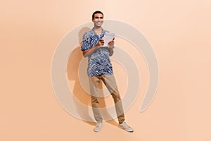 Full size photo of optimistic clever handsome man wear print shirt brown trousers hold tablet in hands isolated on beige