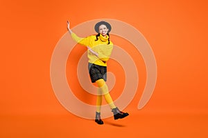Full size photo of optimistic funky girl dance go wear yellow sweater cap skirt shoes isolated on orange background
