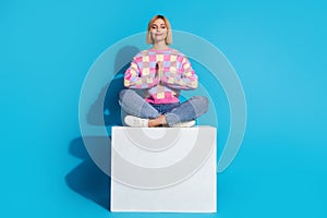 Full size photo of pleasant pretty woman dressed colorful sweater sit on cube hold palms together isolated on blue color