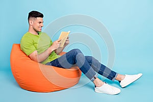 Full size photo of positive young man sit bean bag look read book smile hobby isolated on blue color background