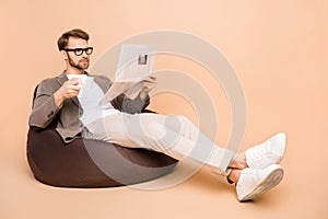 Full size photo of serious focused young man sit chair read paper drink coffee isolated on beige color background