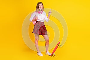 Full size photo of small girl point with skate headphones wear shirt skirt bag sneakers isolated on yellow background