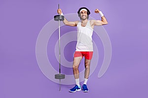 Full size photo of young funky smiling man in glasses brunette hair showing muscles hold barbell isolated on purple photo
