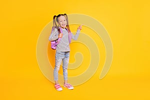 Full size photo of young happy smiling school girl wear backpack pointing finger in copyspace  on yellow color
