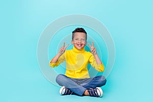 Full size portrait of cheerful positive boy sitting floor demonstrate v-sign isolated on turquoise color background