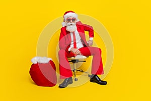 Full size portrait of confident serious aged man sit chair big presents bag isolated on yellow color background