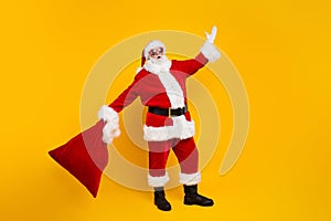 Full size portrait of eccentric overjoyed aged santa hold new year presents bag raise hand isolated on yellow color