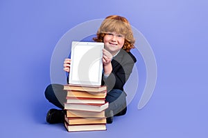 Full size portrait of small boy sit floor pile stack book showing empty space tablet display isolated on purple color