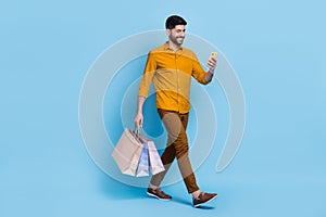 Full size portrait of walking handsome guy use telephone hold packages isolated on blue color background