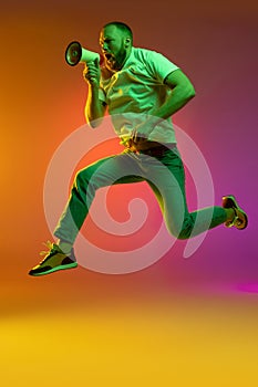 Full size portrait of young caucasian man jumping with megaphone isolated over gradient orange pink background in neon