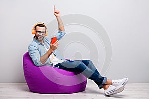 Full size profile photo of funny guy sitting cozy armchair holding telephone earflaps listen youth song raising finger