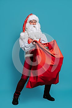 Full size of Surprised stylish Santa Claus holds huge red bag with many presents on Christmas. New Year concept