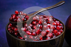 A full wooden bowl with pomegranate seeds on which is a wooden spoon. A bowl full of seeds with a spoonful of seeds on a blue