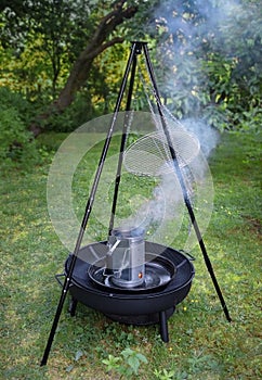 fuming barbecue charcoal chimney starter on a black tripod swivel grill in the garden photo