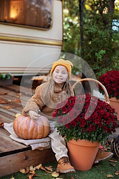 Fun Child in autumn garden with yellow pumpkins and flowers. Happy little girl sitting on porch of house with potted chrysanthemum