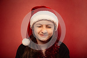 Fun child girl screw up one`s eyes guessing the wish in santa claus hat on red background. Happy Christmas closeup