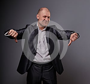 Fun comic bald business man in black suit showing the finger success thumb down sign on grey background. Closeup