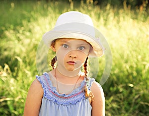 Fun kid girl in fashion hat looking her big opened shocked eyes with humor face on summer green grass background. Closeup