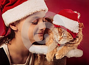 Fun kid girl playing the orange maine coon cat in christmas santa claus cap  on red background. Closeup portrait