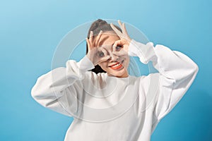Fun and people concept - smiling young woman or teenage girl looking through finger glasses over light blue background