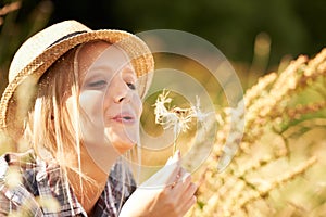 Fun and whimsy in the fields. Beautiful young woman wearing a straw fedora and blowing at a dandelion.