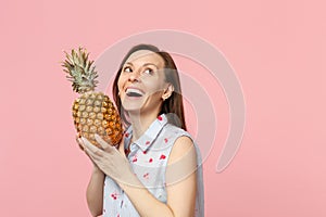 Fun young woman in summer clothes looking up, holding fresh ripe pineapple fruit isolated on pink pastel wall background