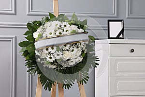 Funeral wreath of flowers on wooden stand and photo frame with black ribbon on white commode indoors