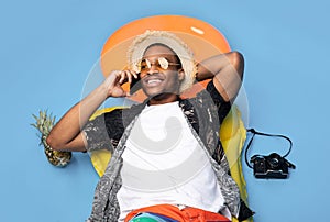 Funky black guy in trendy summer clothes lying on inflatable lilo, speaking on smartphone over blue background, top view