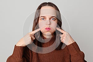 Funky serious brown haired adult woman wearing brown jumper inflate her cheeks pointing index finger showing joke posing isolated