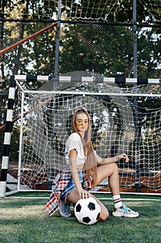 Funky young blonde teenage hipster girl posing with serious face with soccer ball in front of goal post at the stadium