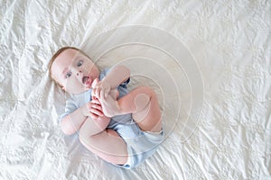 funny 6 months baby boy playing with legs, lying on bed at home on white bedding. top view.