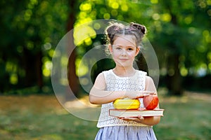 Funny adorable little kid girl with book, apple and backpack on first day to school or nursery. Child outdoors on warm