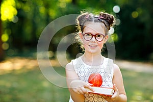 Funny adorable little kid girl with glasses, book, apple and backpack on first day to school or nursery. Child outdoors