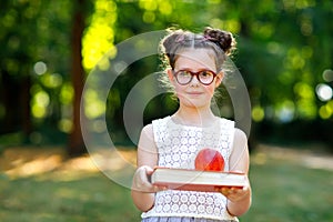 Funny adorable little kid girl with glasses, book, apple and backpack on first day to school or nursery. Child outdoors