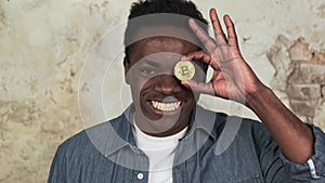 Funny african american man with charming smile standing covering one eye with golden bitcoin close-up. Cryptocurrency