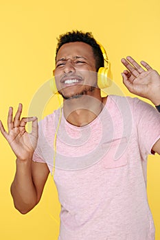Funny african american young man listening music in headphones and dancing over yellow background.