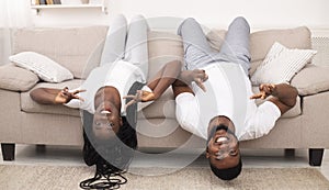 Funny afro couple shoving peace gesture, lying upside-down on sofa