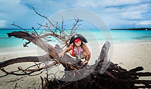 Funny, angry little girl pirate sitting on old dead tree at the beach against dark dramatic sky and ocean background