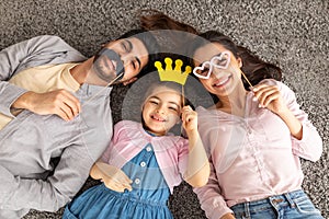 Funny arab father, mother and daughter having fun with crown, glasses and moustache on sticks, lying on carpet, top view